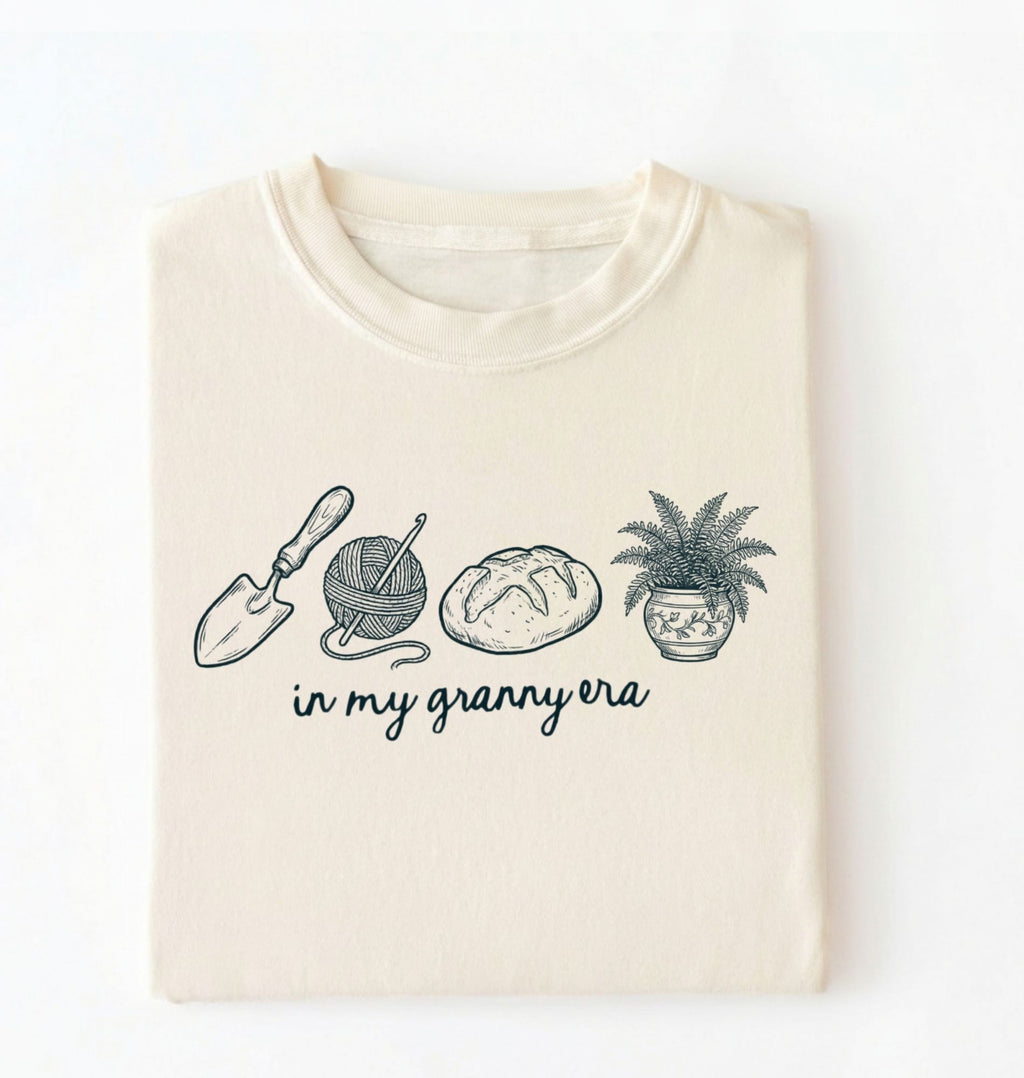 Flat lay of an ivory t-shirt featuring delicate line drawings of gardening tools, yarn, bread, and a plant pot with text reading “in my granny era.”