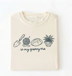 Flat lay of an ivory t-shirt featuring delicate line drawings of gardening tools, yarn, bread, and a plant pot with text reading “in my granny era.”