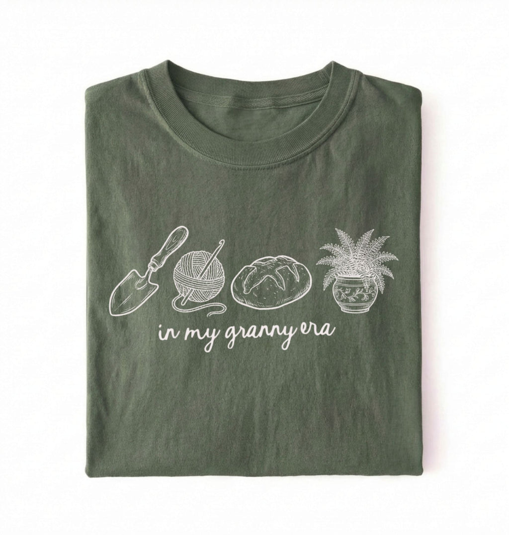 Flat lay of a moss t-shirt featuring delicate line drawings of gardening tools, yarn, bread, and a plant pot with text reading “in my granny era.”