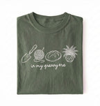 Flat lay of a moss t-shirt featuring delicate line drawings of gardening tools, yarn, bread, and a plant pot with text reading “in my granny era.”