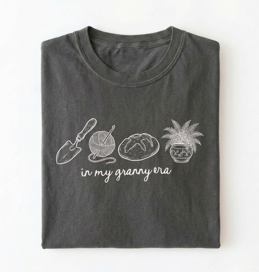 Flat lay of a pepper t-shirt featuring delicate line drawings of gardening tools, yarn, bread, and a plant pot with text reading “in my granny era.” on a white background 