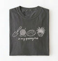 Flat lay of a pepper t-shirt featuring delicate line drawings of gardening tools, yarn, bread, and a plant pot with text reading “in my granny era.” on a white background 