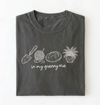 Flat lay of a pepper t-shirt featuring delicate line drawings of gardening tools, yarn, bread, and a plant pot with text reading “in my granny era.” on a white background 