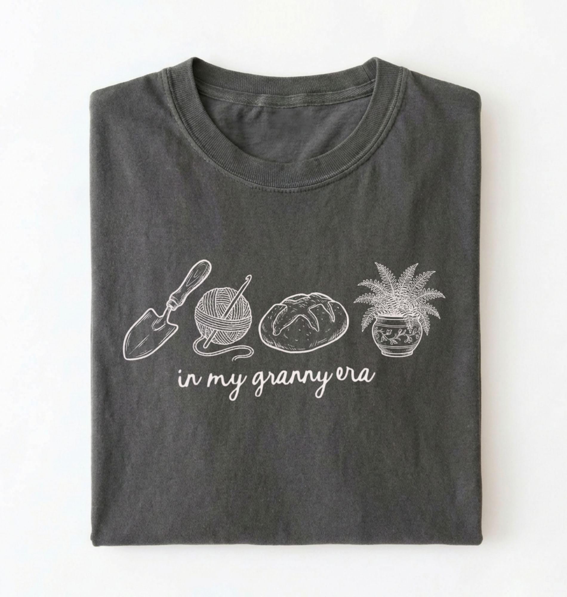 Flat lay of a pepper t-shirt featuring delicate line drawings of gardening tools, yarn, bread, and a plant pot with text reading “in my granny era.” on a white background 