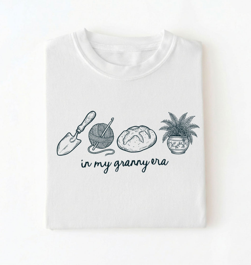 Flat lay of a white t-shirt featuring delicate line drawings of gardening tools, yarn, bread, and a plant pot with text reading “in my granny era.”