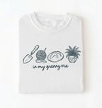 Flat lay of a white t-shirt featuring delicate line drawings of gardening tools, yarn, bread, and a plant pot with text reading “in my granny era.”