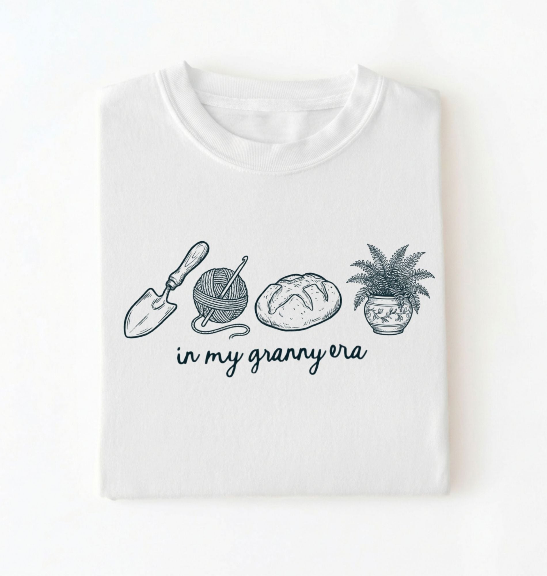 Flat lay of a white t-shirt featuring delicate line drawings of gardening tools, yarn, bread, and a plant pot with text reading “in my granny era.”