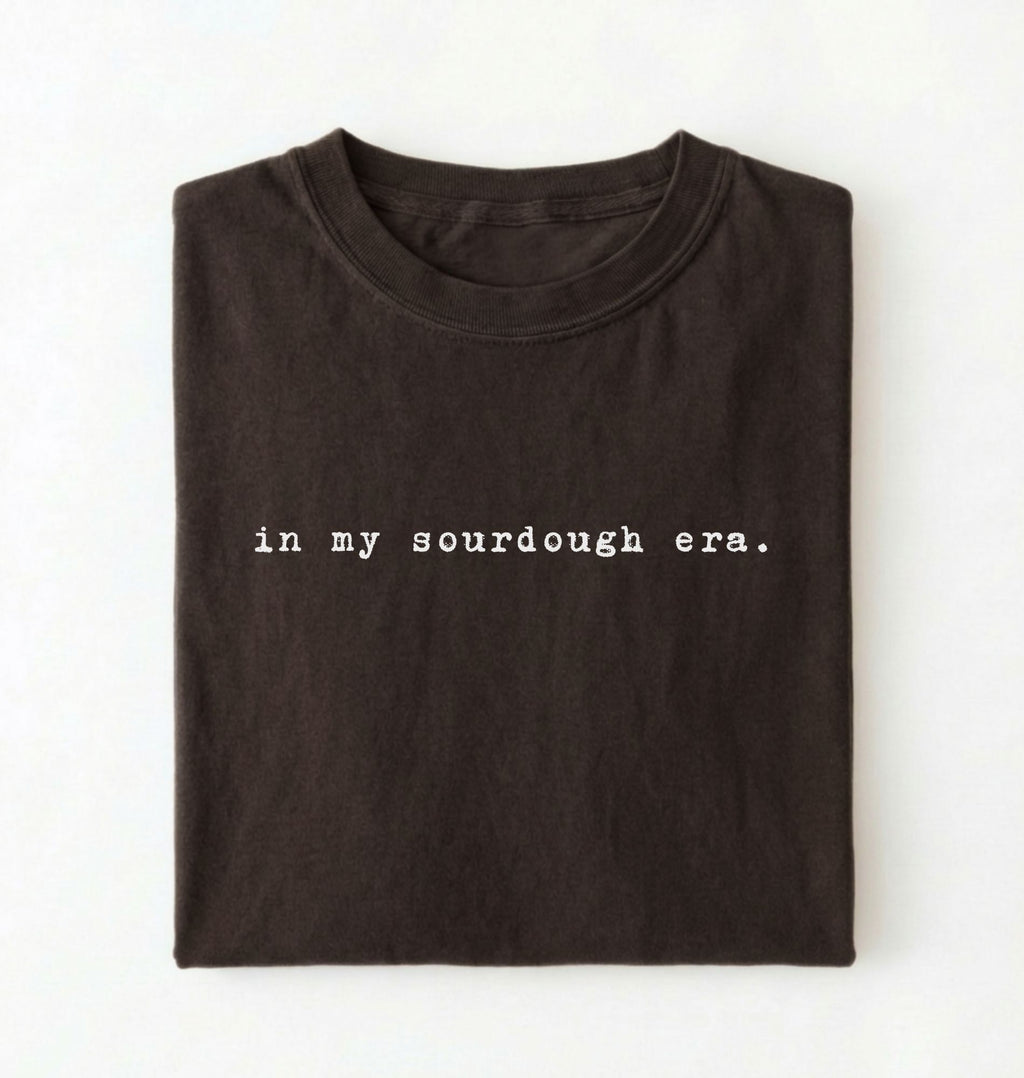 Flat lay of a dark chocolate Funny in my sourdough era baking shirt perfect for bread lovers home bakers and cozy kitchen aesthetic on a white background 