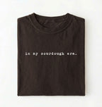 Flat lay of a dark chocolate Funny in my sourdough era baking shirt perfect for bread lovers home bakers and cozy kitchen aesthetic on a white background 
