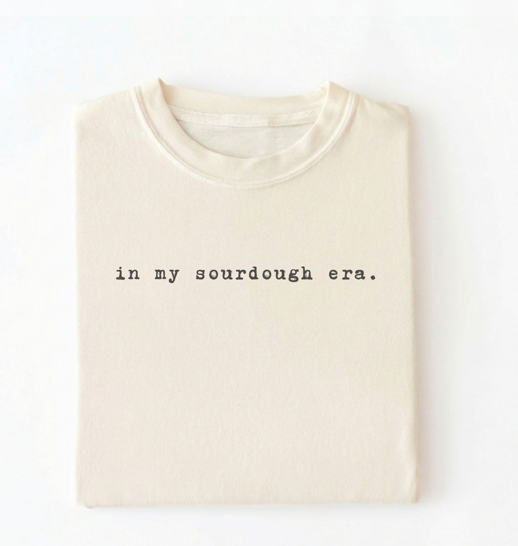 Flat lay of a ivory Funny in my sourdough era baking shirt perfect for bread lovers home bakers and cozy kitchen aesthetic on a white background
