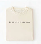 Flat lay of a ivory Funny in my sourdough era baking shirt perfect for bread lovers home bakers and cozy kitchen aesthetic on a white background