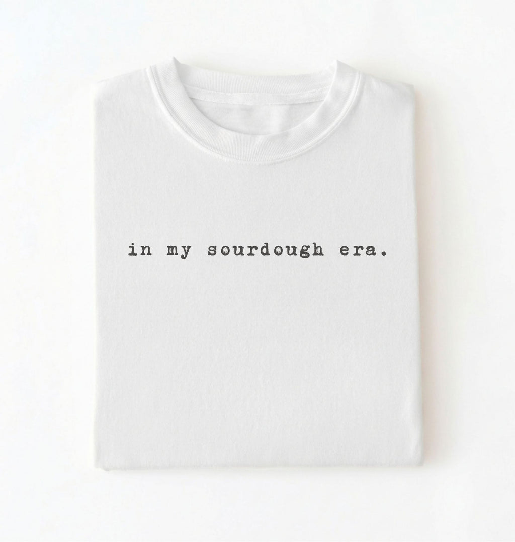 Flat lay of a white Funny in my sourdough era baking shirt perfect for bread lovers home bakers and cozy kitchen aesthetic on a white background