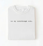 Flat lay of a white Funny in my sourdough era baking shirt perfect for bread lovers home bakers and cozy kitchen aesthetic on a white background
