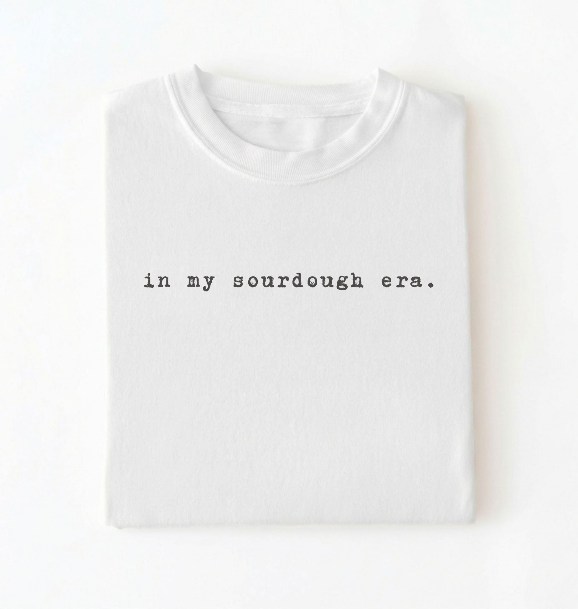 Flat lay of a white Funny in my sourdough era baking shirt perfect for bread lovers home bakers and cozy kitchen aesthetic on a white background