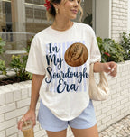 Woman wearing a t-shirt with 'In My Sourdough Era' design, holding a drink, standing outdoors.