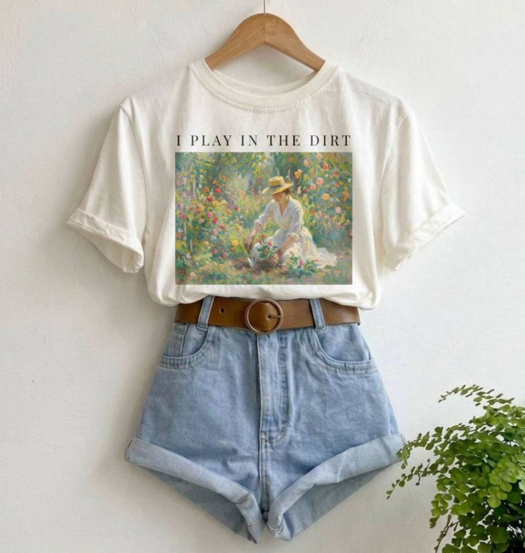 an image of a hanging white shirt with a vintage washed out design of a woman in the garden that says "I play in the dirt"