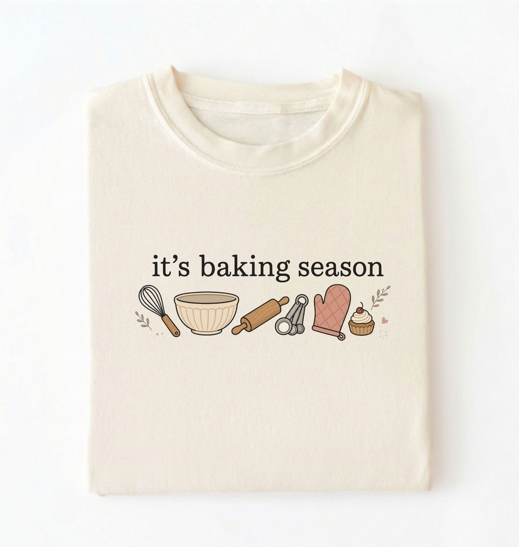 Flat lay of a ivory its baking season shirt with baking tools cupcake and oven mitt cute baker gift on a white background 