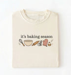 Flat lay of a ivory its baking season shirt with baking tools cupcake and oven mitt cute baker gift on a white background 
