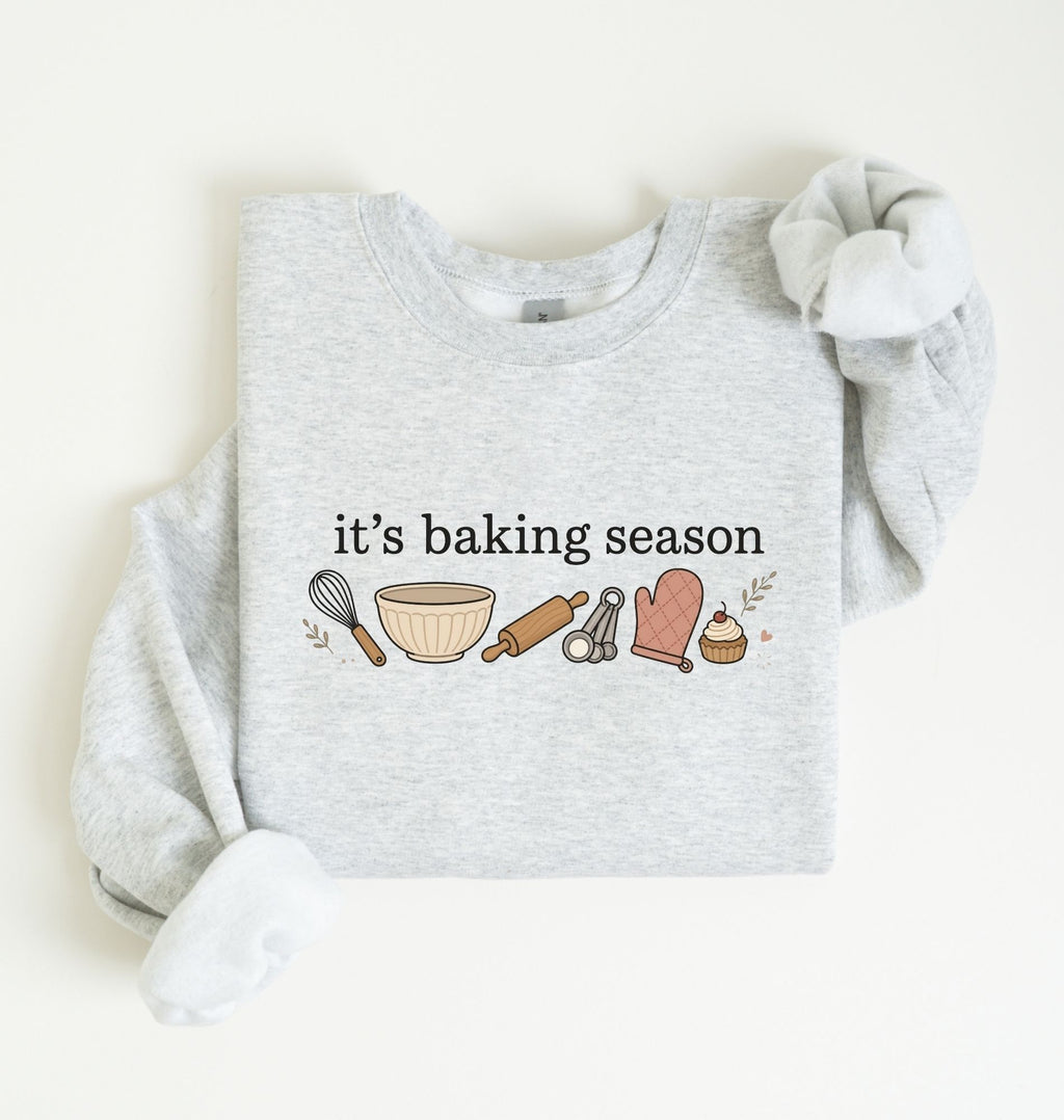 Flat lay of an ash It's Baking Season graphic sweatshirt featuring baking tools and cupcake design on a white background 