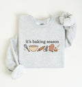 Flat lay of an ash It's Baking Season graphic sweatshirt featuring baking tools and cupcake design on a white background 