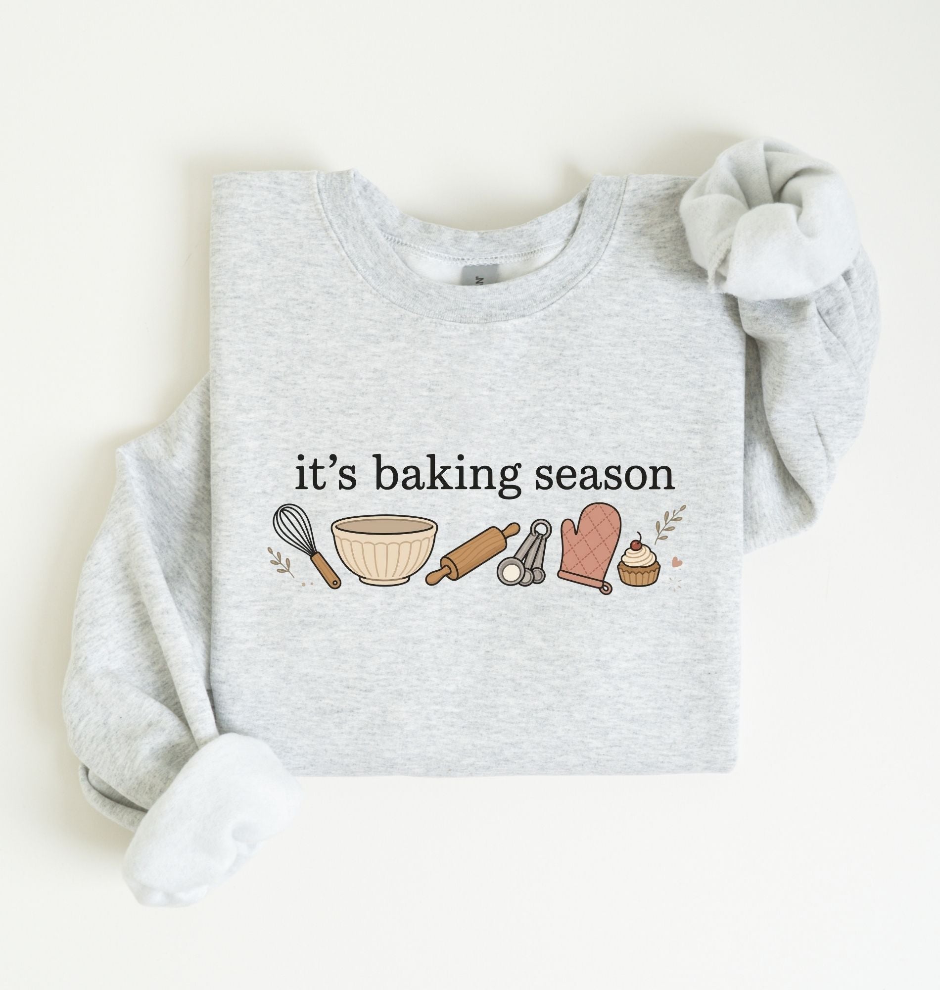 Flat lay of an ash It's Baking Season graphic sweatshirt featuring baking tools and cupcake design on a white background 