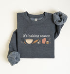 Flat lay of a dark heather It's Baking Season graphic sweatshirt featuring baking tools and cupcake design on a white background 