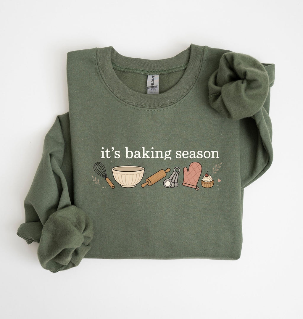 Flat lay of a green It's Baking Season graphic sweatshirt featuring baking tools and cupcake design on a white background 