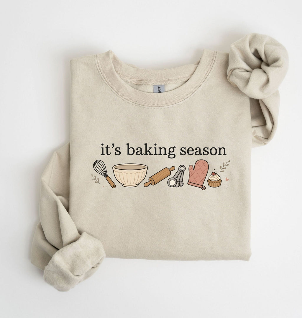 Flat lay of a sand It's Baking Season graphic sweatshirt featuring baking tools and cupcake design on a white background 