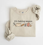 Flat lay of a sand It's Baking Season graphic sweatshirt featuring baking tools and cupcake design on a white background 