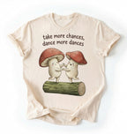 Flat lay of a Natural t-shirt with two dancing mushrooms and the phrase take more chances dance more dances, whimsical cottagecore gift tee on a white background 