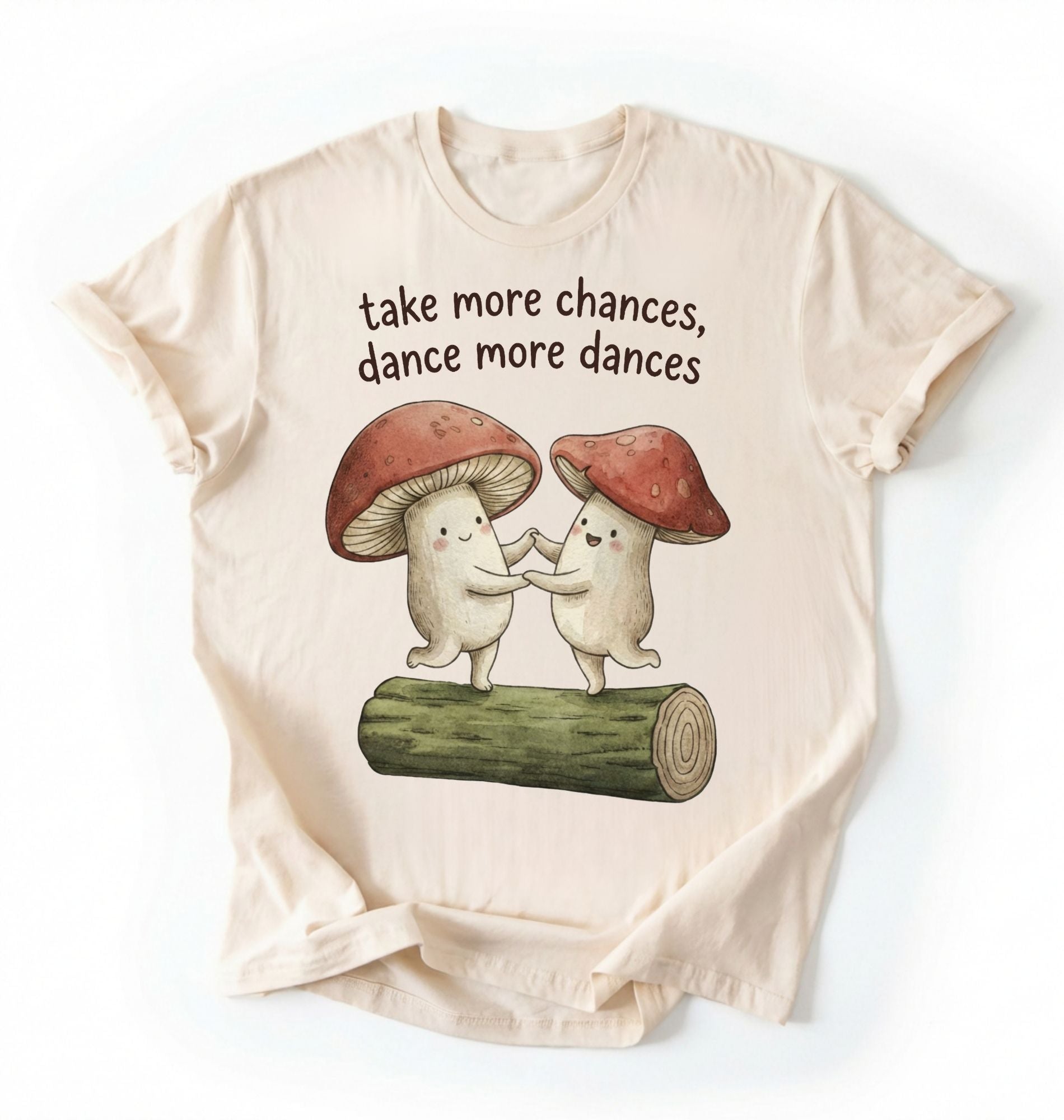 Flat lay of a Natural t-shirt with two dancing mushrooms and the phrase take more chances dance more dances, whimsical cottagecore gift tee on a white background 