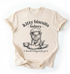 Flat lay of an ivory Kitty Biscuits Bakery shirt featuring a baking kitten and dough on cream t-shirt on  a white background 