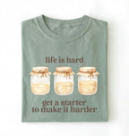 Flat lay of a bay Funny Life Is Hard Get a Starter sourdough shirt with sourdough starter jars on brown t-shirt on a white background 
