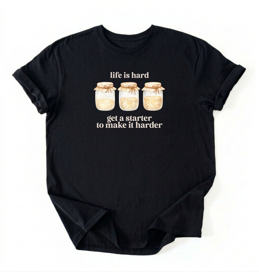 Flat lay of a black Funny Life Is Hard Get a Starter sourdough shirt with sourdough starter jars on brown t-shirt on a white background 