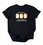 Flat lay of a black Funny Life Is Hard Get a Starter sourdough shirt with sourdough starter jars on brown t-shirt on a white background 