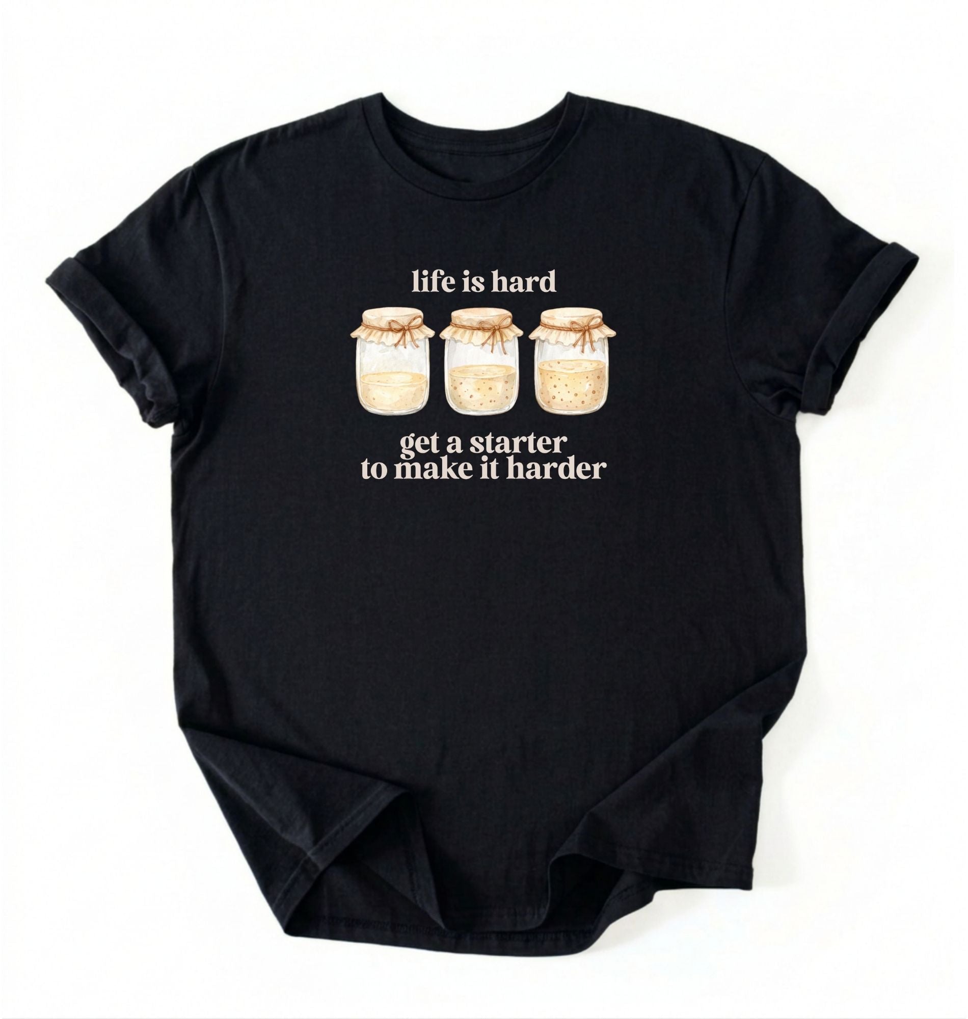 Flat lay of a black Funny Life Is Hard Get a Starter sourdough shirt with sourdough starter jars on brown t-shirt on a white background 