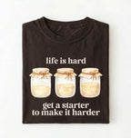 Flat lay of a brown Funny Life Is Hard Get a Starter sourdough shirt with sourdough starter jars on brown t-shirt on a white background 