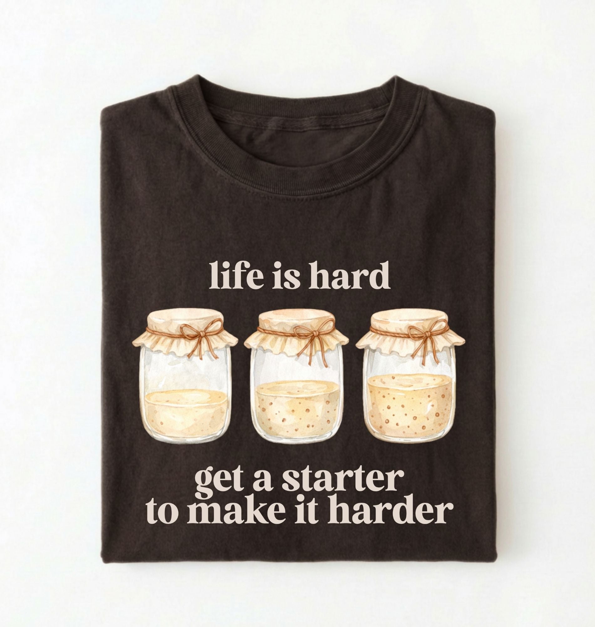 Flat lay of a brown Funny Life Is Hard Get a Starter sourdough shirt with sourdough starter jars on brown t-shirt on a white background 