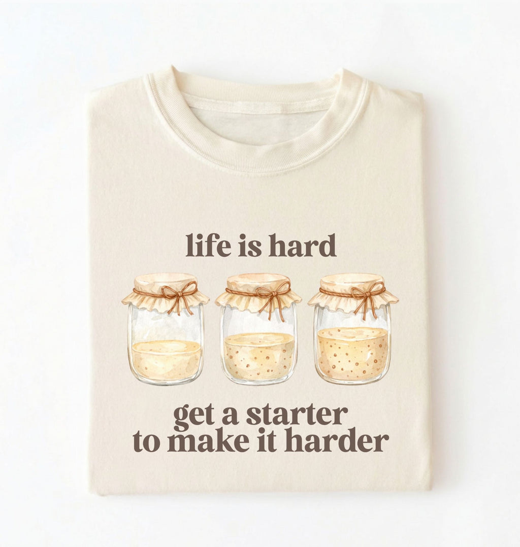 Flat lay of a cream Funny Life Is Hard Get a Starter sourdough shirt with sourdough starter jars on brown t-shirt on a white background 