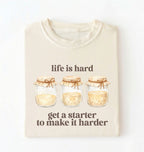 Flat lay of a cream Funny Life Is Hard Get a Starter sourdough shirt with sourdough starter jars on brown t-shirt on a white background 