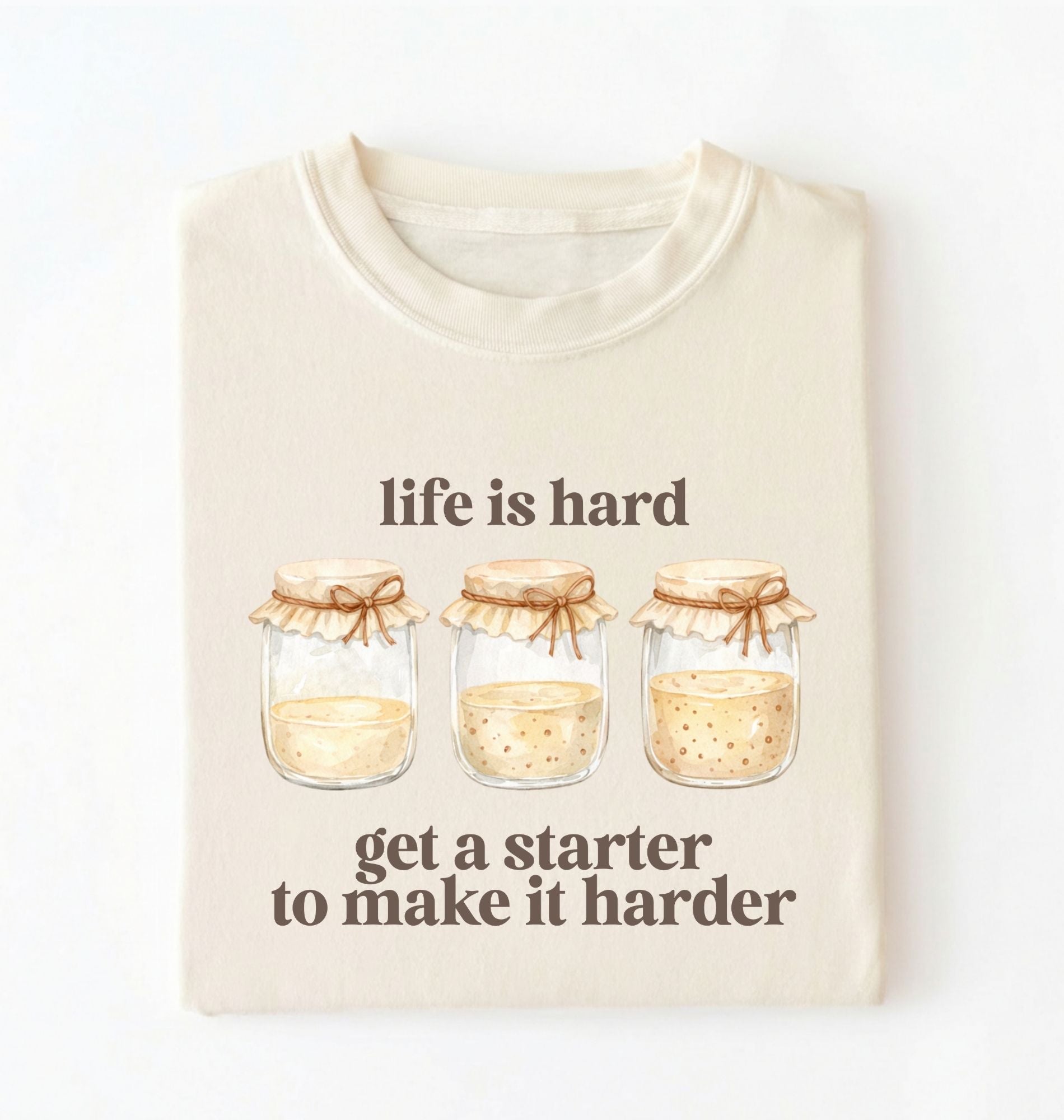 Flat lay of a cream Funny Life Is Hard Get a Starter sourdough shirt with sourdough starter jars on brown t-shirt on a white background 