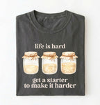 Flat lay of a pepper Funny Life Is Hard Get a Starter sourdough shirt with sourdough starter jars on brown t-shirt on a white background 