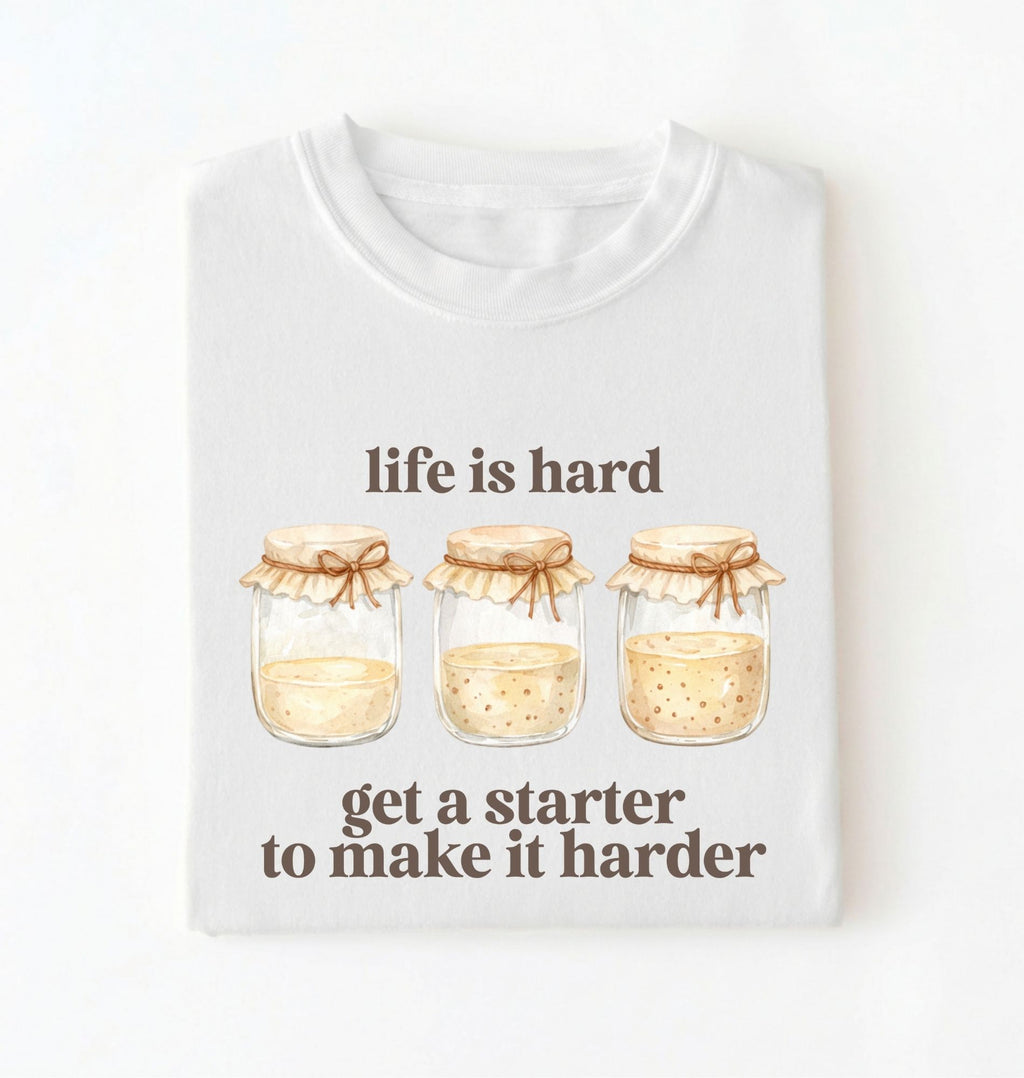 Flat lay of a white Funny Life Is Hard Get a Starter sourdough shirt with sourdough starter jars on brown t-shirt on a white background 