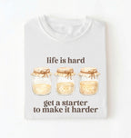 Flat lay of a white Funny Life Is Hard Get a Starter sourdough shirt with sourdough starter jars on brown t-shirt on a white background 
