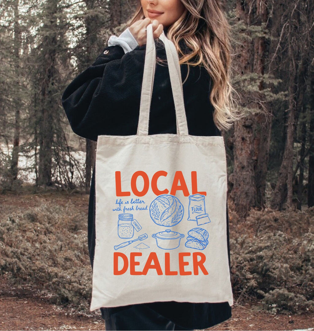 Person holding a tote bag with 'Local Dealer' text and graphics in a forest setting