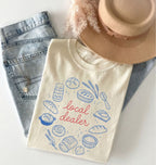Beige t-shirt with bread illustrations and 'local dealer' text, placed on denim shorts with a hat on top.