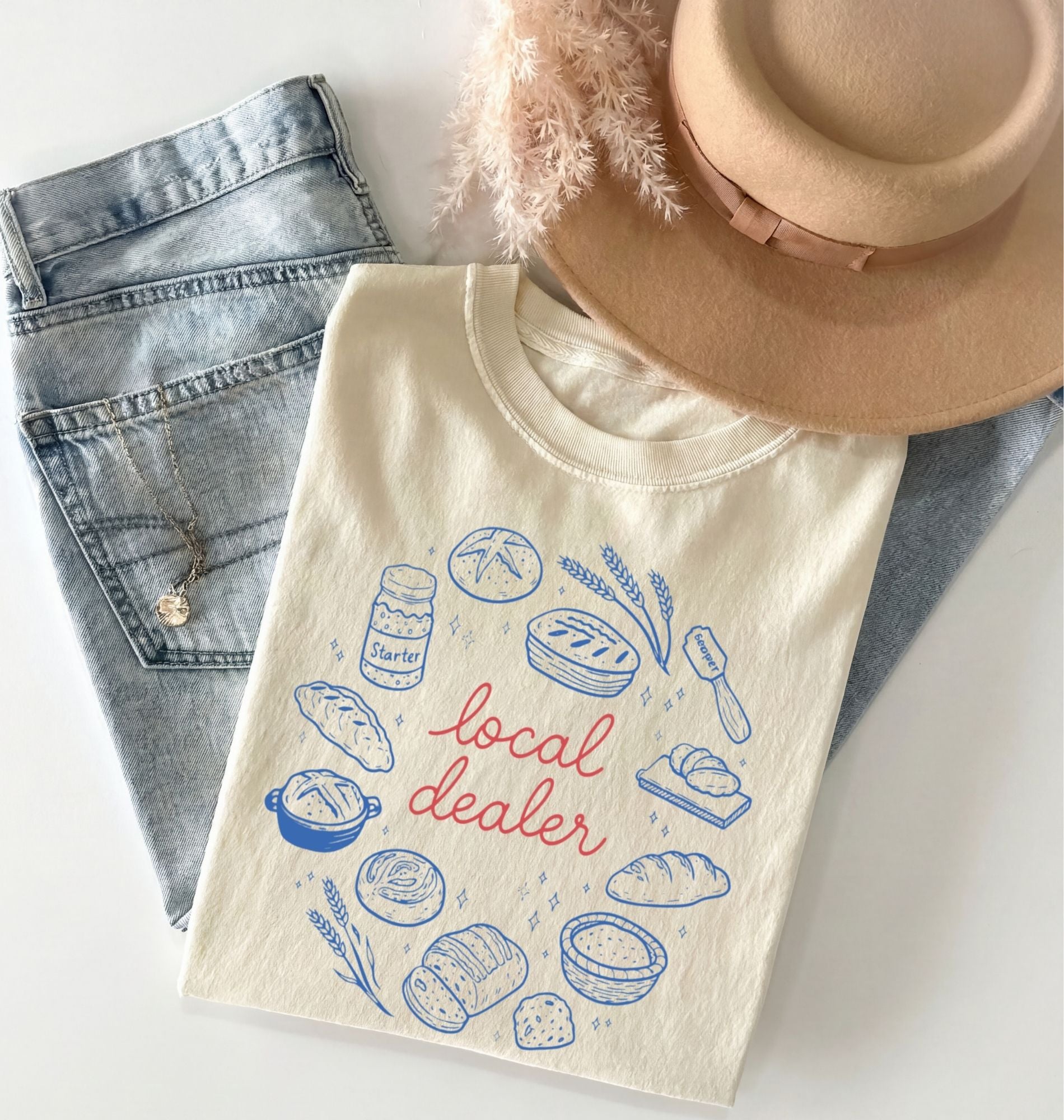 Beige t-shirt with bread illustrations and 'local dealer' text, placed on denim shorts with a hat on top.