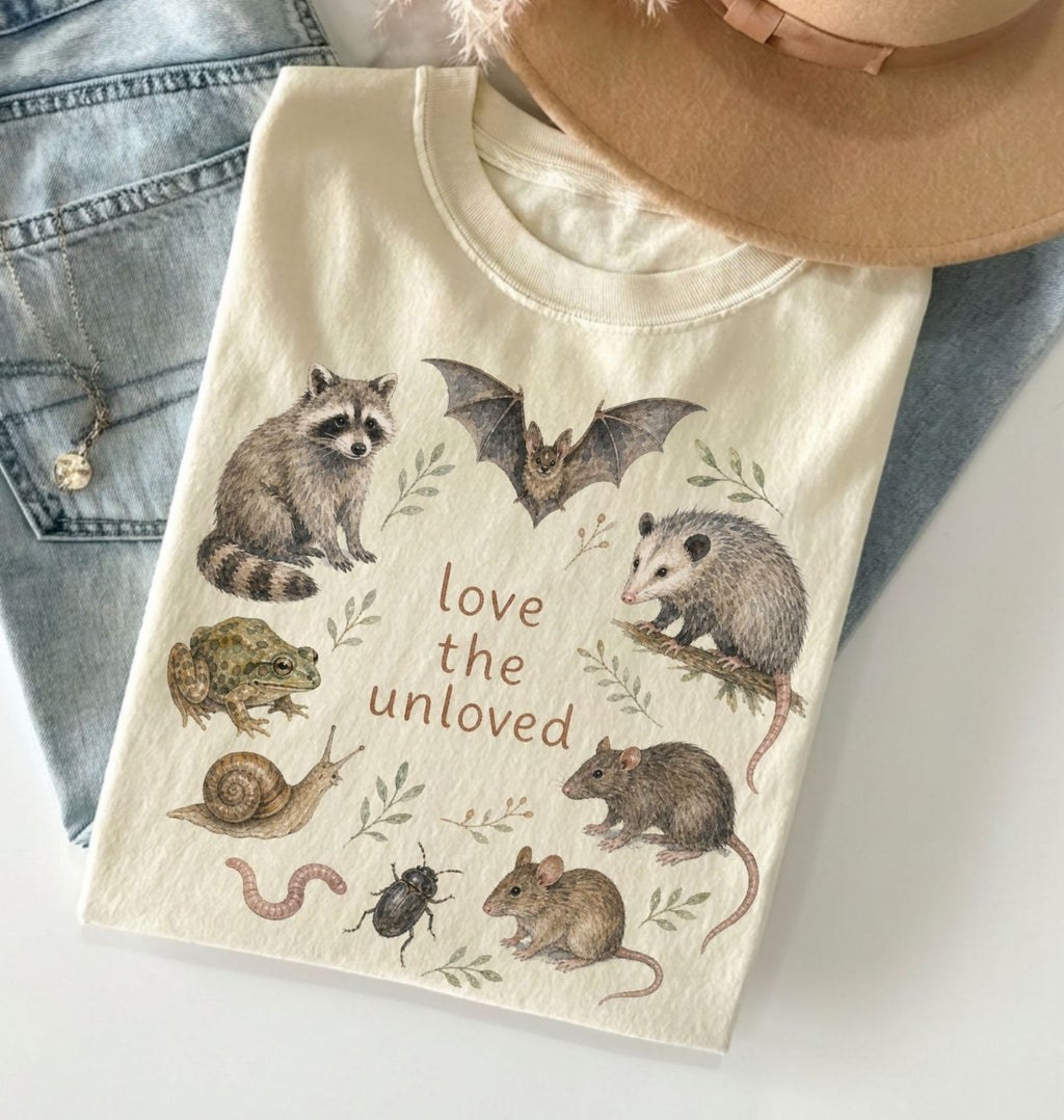 Beige t-shirt with animal illustrations and 'love the unloved' text, displayed on a light blue denim surface.