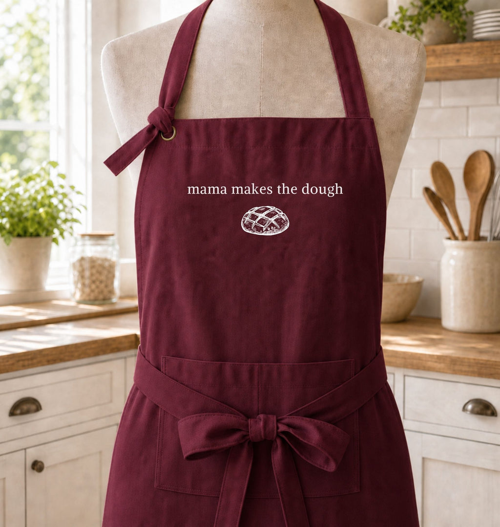 A burgundy kitchen apron displayed on a mannequin torso in a bright, white-tiled kitchen. The apron features the white embroidered text 'mama makes the dough' and a small white bread graphic on the chest, along with a matching waist tie and front pocket.