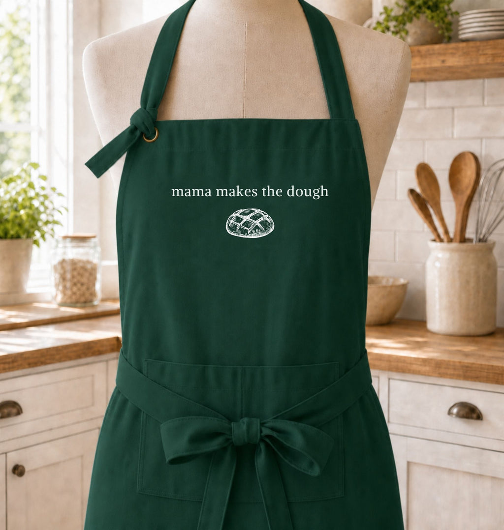 A dark green kitchen apron displayed on a mannequin torso in a bright kitchen setting. The apron has white text reading 'mama makes the dough' above a small white bread graphic, and a waist tie with a bow.